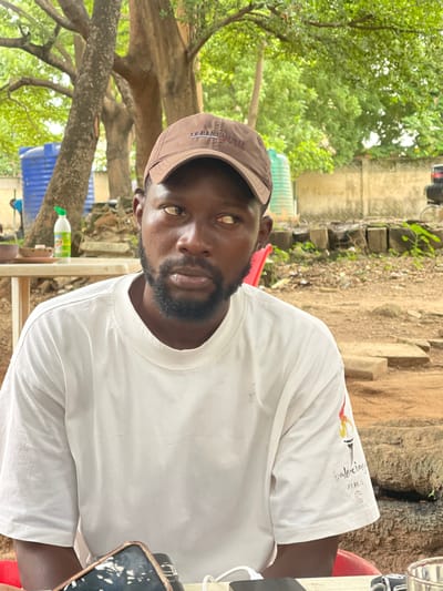 A man is captured in a medium shot, seated outdoors with a blurred background of trees and a concrete wall. He wears a brown baseball cap and a white t-shirt with a graphic design on the right sleeve featuring a torch and the text "Balenciaga Paris." He has dark skin, a beard, and his eyes are looking to his left, away from the camera. A white plastic bottle with a green cap sits on a table to his left, along with a shallow bowl. A portion of a phone is visible on the table in front of him, and a glass is to his right. The environment suggests a casual outdoor setting, possibly a restaurant or gathering area, in Wupa, Nigeria.