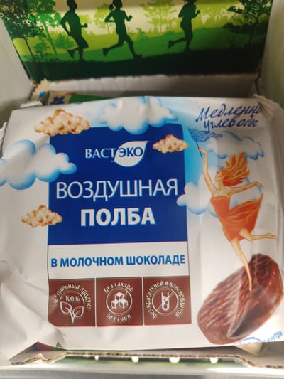 The image shows a close-up view of the packaging for a food product. The packaging is predominantly white and blue, with graphics of clouds and stylized depictions of people running. Text on the packaging is in Russian. One prominent text reads "ВОЗДУШНАЯ ПОЛБА," which translates to "Air Spelt." Below this, it states "В МОЛОЧНОМ ШОКОЛАДЕ," meaning "In Milk Chocolate." There are also circular emblems indicating "100% natural product," "sugar-free," "salt-free," and "free of dyes and preservatives." A graphic of a woman in an orange dress reaching up with a net is also present, as is a depiction of a chocolate-covered food item. The background of the packaging features a silhouette of people running through a green, tree-filled landscape. The overall impression is that of a healthy snack product. There is no information or visual cues in the image to suggest its location in the city of Svobodny, Russia.