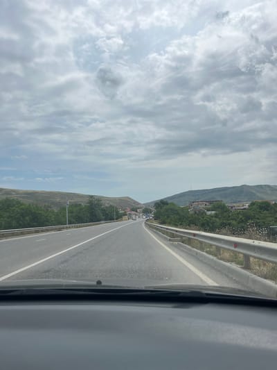The image depicts a view from inside a vehicle driving on a paved road. The road has two lanes in each direction, separated by white lines. A metal guardrail runs along the right side of the road. On the left side of the road, there are green trees and rolling hills. On the right side, there are more trees, some houses, and buildings. In the distance, there are more hills and mountains under a cloudy sky. A small settlement or town can be seen nestled among the hills in the distance. There are no visible people in the image. The scene appears to be a rural or semi-urban setting on a cloudy day. The location is identified as Karabudakhkent, Russia.