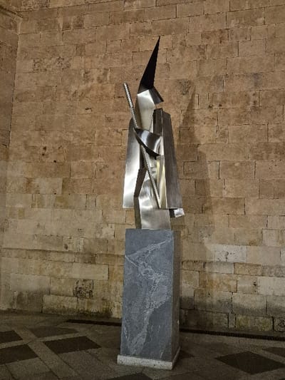 The image displays an abstract, geometric sculpture located in the city of Salamanca, Spain. The sculpture, composed of polished, silver-toned metallic sheets, features sharp angles, intersecting planes, and is topped by a distinct dark, conical finial. It rests on a rectangular pedestal of dark grey, mottled stone with a lighter, slightly wider base. The background consists of a wall constructed from light brown, roughly cut ashlar stone blocks. The ground exhibits a patterned pavement composed of alternating light and dark square tiles, with a narrow linear drainage grate visible adjacent to the wall's base. The scene is illuminated, likely under evening or night conditions, casting a prominent shadow of the sculpture onto the stone wall to its right.