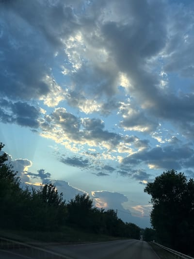 A twilight scene unfolds with a road cutting through a dark, tree-lined landscape under a dramatic sky. The sky is a canvas of blues and grays, with pockets of golden sunlight breaking through scattered clouds, casting ethereal rays. The foreground features the asphalt of a road, with a guardrail on the right, and dark, dense foliage lining both sides. The trees are silhouetted against the brightening sky, suggesting a transition from day to night. In the distance, the road curves slightly, and a few faint lights are visible, hinting at the presence of vehicles or distant structures. The overall atmosphere is serene and contemplative, with the natural beauty of the sky dominating the composition. The image is set in Neznamovo, Russia, and captures a moment of natural splendor.