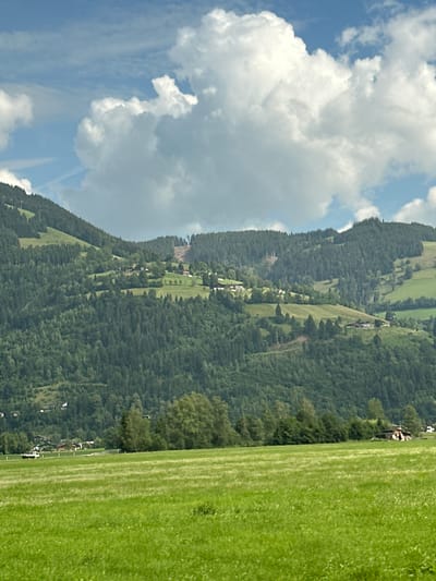 The image depicts a verdant, rolling landscape under a partly cloudy sky. In the foreground, a vast field of bright green grass stretches across the frame. Beyond the field, the middle ground is dominated by a dense forest of dark green trees that climb the slopes of several layered mountains. Scattered throughout the mountainside are small clusters of buildings, appearing to be houses or chalets, with some rooftops visible amidst the trees and on the green, cleared patches of the slopes. A visible path or road, possibly a ski slope due to its cleared nature, ascends one of the mountains in the distance. The sky above is a mix of bright blue and large, fluffy white clouds. In the far left of the middle ground, a white vehicle is parked on the edge of the field. The scene is set in the vicinity of Bruck an der Großglocknerstraße, Austria, a region known for its mountainous terrain and picturesque villages.