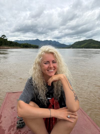 In Thinhong, Laos, a woman with long, curly blonde hair sits on the edge of a red boat. She is wearing a grey t-shirt and has a tattoo on her right arm. Her left hand rests on her knee, and her right hand is supporting her chin as she smiles at the camera. The boat is on a wide, muddy river. In the background, lush green hills and mountains rise under a cloudy sky. The scene depicts a serene moment during a boat trip on the river.