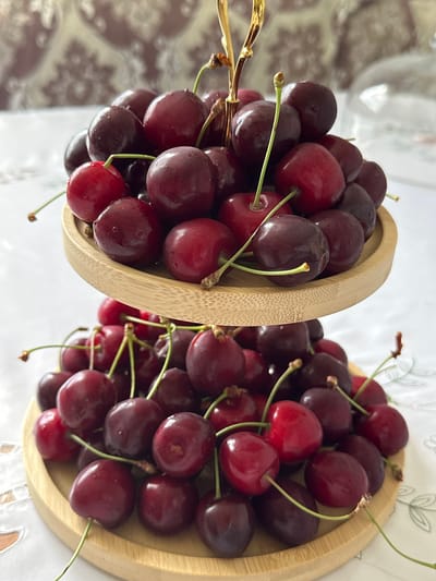 A two-tiered bamboo serving tray holds a generous amount of dark red cherries. The tray is positioned on a white tablecloth with a subtle embroidered pattern. The background is out of focus, showing a textured wallpaper and a clear glass object. The lighting is bright, creating highlights on the glossy cherry skins and indicating an indoor setting, likely a home or event space, in Кашат, Kyrgyzstan. There are no people visible in the image.