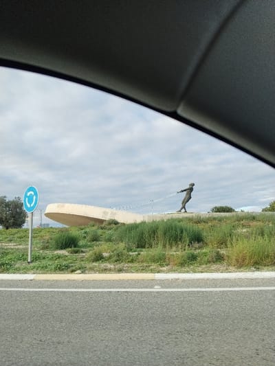 The image, captured from inside a vehicle, displays a public sculpture within a roadside environment under an overcast sky in San Javier, Spain. The upper portion of the frame is occupied by the dark gray interior trim of the vehicle.

In the foreground, a segment of asphalt road is visible, featuring a solid white lane line and a yellow curb to its right. Beyond the road, an unkempt grassy embankment covered with varying heights of green and dry vegetation ascends.

Positioned on this embankment, towards the center-right, is a dark-colored, anthropomorphic sculpture of a person, appearing to be male, in a straining posture. This figure pulls a metallic chain that extends leftwards towards a large, light-colored concrete structure. This structure, resembling a stylized boat hull or wave, is partially embedded in the embankment behind the figure and curves upwards.

To the left of the scene, on a metal pole emerging from the grassy area, a standard blue circular road sign with white arrows indicating a roundabout is visible. The sky is predominantly gray and cloudy. Minimal distant foliage is discernible on the horizon to the far left and right.