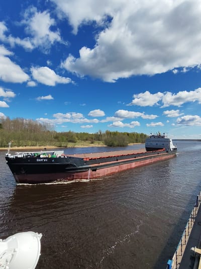The image shows a large barge, named "Vaygach," moving on a river, likely in the vicinity of the city of Pruzhinino, Russia based on the name of the ship, which is the name of a territory in Russia. The barge is dark in color, likely red and black, and the surrounding trees suggest a riverbank. The sky is a bright blue with scattered white clouds. The water is dark and reflects some of the sky's color. The overall scene suggests a sunny day, perhaps during a period of foliage growth along the river. The barge is oriented to the right of the frame, with its front (bow) visible. Parts of the other banks of the river are visible.