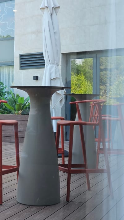 This media file shows an outdoor patio with a table and chairs, possibly in Alhama de Aragón, Spain. The scene is oriented vertically. A large, light-colored umbrella is partially open and lies across a table with a unique, hourglass-shaped base. The table's top is not fully visible, but its base is dark gray. Red chairs, with a modern, angular design, surround the table and are reflected in a large window or glass panel behind them. The patio floor is made of wooden planks. Through the glass, lush green trees and foliage are visible, suggesting a pleasant outdoor setting, likely during the daytime with good weather. The overall impression is one of a relaxed, outdoor dining or lounge area. There is no visible text in the image.