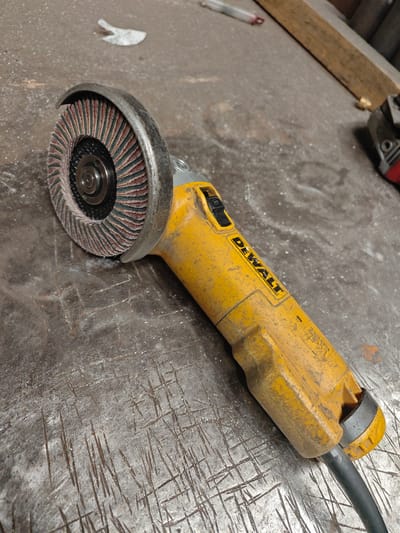 An angle grinder with a flap disc is lying on a scratched metal surface. The angle grinder is yellow with black accents and the brand name "DEWALT" is visible on its body. The flap disc is composed of layered abrasive material in red and green. The metal surface appears to be a workbench or similar industrial setting. There are no people or any other objects clearly identifiable in the immediate vicinity, though other tools or equipment are faintly visible in the blurred background. The scene is indoors, and the lighting suggests it may be in a workshop. The city of Boulogne-sur-Mer, France, is not depicted in the image.