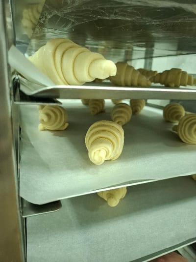 The image displays unbaked dough products arranged within a multi-tiered metallic rack, located in Bnei Dror, Israel. The rack holds several baking trays, each lined with white parchment paper. On the uppermost visible tray, a single large, raw, crescent-shaped croissant is positioned in the foreground, with multiple smaller, unbaked croissants visible further back on the same level. The middle tray contains numerous small, spiral-shaped raw dough pieces scattered across its surface. A lower tray with parchment paper and a partially visible small dough item is also present at the bottom of the frame. The top of the rack is covered with clear plastic film, exhibiting creases and reflections. The scene is brightly illuminated, indicating an indoor commercial food preparation environment. No individuals are visible.