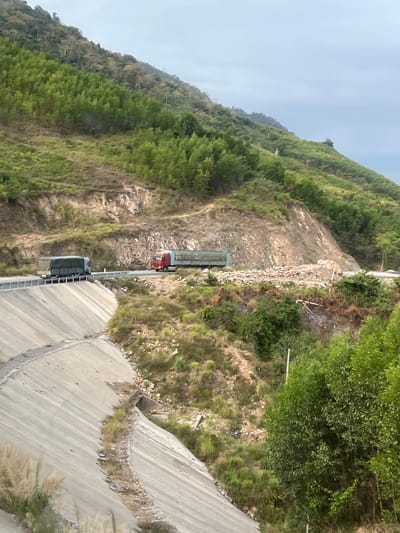 The image depicts a mountainous landscape in Aê Thi, Vietnam, showcasing a road carved into the hillside. A red semi-truck is parked on the road, while a grey semi-truck is further down the road. The roadside features rocky terrain and vegetation. Concrete infrastructure flanks the road, including what appears to be drainage systems. The backdrop is a steep, tree-covered mountain, and the sky is overcast.