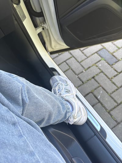 A photograph taken from inside a car shows a person's leg resting on the car door. The person is wearing light blue jeans and a white sneaker, specifically a New Balance 530 model. The car door is open, revealing a view of a brick sidewalk or street outside. A car door hinge and an information label are visible on the car frame. The location is inferred to be in Wassenberg, Germany based on the prompt's requirements.