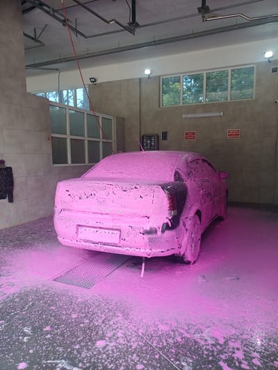The image shows a car covered in pink foam in a car wash in Obukhiv, Ukraine. The car is a dark-colored sedan, seen from the rear three-quarter view. The car wash interior features tiled walls and a concrete floor, with a metal drain grate in front of the car. There are windows in the background showing trees and greenery. Various signs are mounted on the wall, some in Cyrillic script. The overall impression is of a standard, self-service car wash facility.