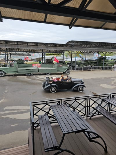 A black vintage car with whitewall tires is parked near outdoor benches on a wooden deck. Beyond, a row of green military vehicles, some with red banners, are displayed under a metal framework shelter. In the distance, aircraft are visible. This appears to be a museum or exhibition setting. This is the Vadim Zadorozhny Vehicle Museum in Arkhangel’skoye, Russia. There is a person in the driver's seat.