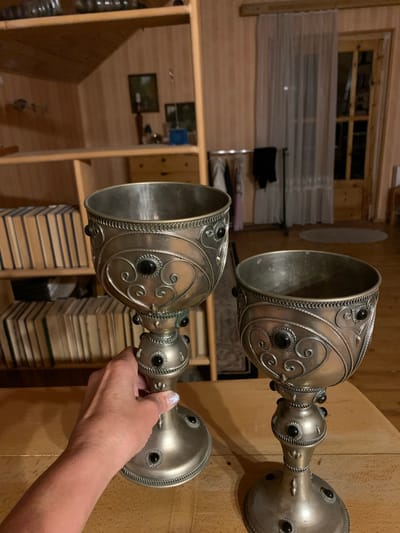 The image shows two ornate, silver goblets with black stone accents, placed on a wooden surface. One goblet is held by a woman's hand, which is visible in the foreground. The goblets have intricate scrollwork and beaded edges. In the background, wooden shelves are filled with stacks of books. There is also a window with a view of what appears to be a rural or rustic setting, with wooden walls and structures visible. The lighting suggests an indoor environment, possibly during the day. The scene conveys a sense of history or tradition, with the goblets appearing to be antique or decorative items. There is no visible text in the image. The location context provided is Mepiskalaki, Georgia.