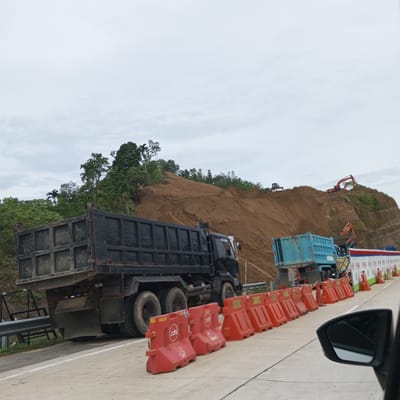 The image shows a construction site on a hillside in Lonbaroh, Indonesia. A large black dump truck is parked on the side of a paved road, facing away from the viewer. In the background, the hillside is covered in loose brown soil, with evidence of excavation and earthmoving. There are several pieces of heavy machinery visible on the hillside, including an orange excavator and a white bulldozer. A light blue dump truck is also present further down the road. The area is cordoned off with red and orange traffic barriers. The sky is overcast.