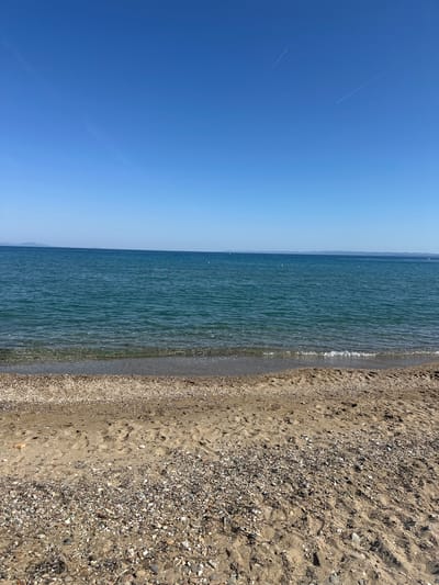 The image depicts a coastal scene in Kalyves Polygyrou, Greece. A strip of sandy beach mixed with pebbles occupies the right side of the frame. Gentle waves are lapping at the shoreline. The sea, ranging in color from light to dark blue, extends across the middle portion of the image. The clear blue sky dominates the left side of the frame, with visible contrails of airplanes.
