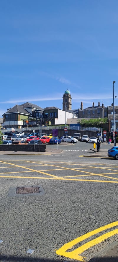 In Enniskillen, United Kingdom, a scene unfolds on a clear, sunny day, featuring a street intersection with prominent yellow box junction markings. In the foreground, the asphalt is textured with a distinct pattern, interrupted by a metal manhole cover and several reflective road studs. A lone person, dressed in dark clothing, stands near a yellow barrier in the middle ground, appearing to be in motion.
Beyond the intersection, a parking lot is filled with various vehicles, including white, blue, red, and yellow cars, as well as a dark-colored SUV. A low stone wall separates the parking lot from a raised area where a building complex stands. This complex features a multi-story structure with a dark roof, a green facade, and a prominent "GUINNESS" sign. To the right of this building, a tall, square tower with a green, domed roof and visible clock faces rises against the bright blue sky, suggesting a landmark or public building. Further to the right, residential-style buildings with brick chimneys are visible.
On the street, traffic signals are present, with one showing a red light. Other signage includes "ONE WAY" arrows and a red circular sign with a diagonal line, indicating "NO ENTRY." A blue car is parked on the right side of the street. The overall environment suggests an urban setting with commercial and residential elements, under a cloudless blue sky.