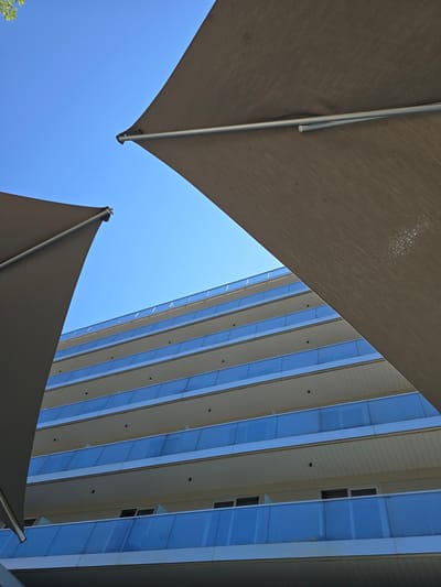 The image is a low-angle shot of a modern building in Rimini, Italy, viewed from beneath two large, dark fabric awnings. The awnings dominate the foreground, with one on the left and another on the right, both angled upwards towards the bright blue sky. The building behind them is a multi-story structure with repeating balconies, each featuring a light-colored facade and a bright blue glass railing. The balconies are stacked in neat horizontal rows, suggesting a hotel or residential building. The sky is clear and cloudless, indicating a sunny day. There are no people visible in the scene. The composition creates a sense of geometric repetition and contrasts the organic shape of the awnings with the rigid lines of the architecture.