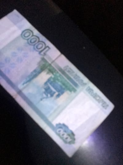 A slightly angled, out-of-focus image reveals a Russian 1000 Ruble banknote. The denomination "1000" is visible in two locations, along with partial images of the banknote's design, including light blue and green colors. The background appears to be a dark, indistinct surface. The top right corner is very blurry.
