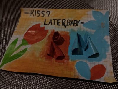 The image shows a handmade drawing on graph paper, featuring colorful watercolor illustrations of flowers. In the top left corner, a red tulip with green leaves is depicted. To the right of the tulip, there are three origami-like folded shapes, one red and two blue, placed on a background of watercolor splashes in orange and blue. Above these elements, the words "-KISS?" are written in black marker, followed by "LATERBABY -". The drawing is placed on a textured, dark grey surface, likely a table or placemat. There is no discernible information within the image that could indicate a location in Dymer, Ukraine. The image is a close-up of the drawing and its immediate surroundings, with no wider environmental context visible.