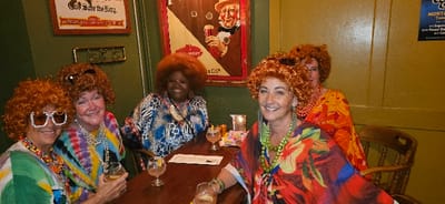 The image depicts an indoor scene within an establishment, presumed to be located in Centralia, United States. Five adult females are gathered around a dark-stained wooden rectangular table. All five individuals are wearing large, curly, reddish-brown wigs and are smiling at the camera, engaged in a social interaction.
From left to right:
1. A light-complexioned female wears a multi-colored patterned top, multiple colorful beaded necklaces, and large white-framed sunglasses over her wig. She holds a clear glass containing a light beverage.
2. A light-complexioned female wears a tie-dye shirt, multiple silver-toned necklaces, and has dark sunglasses perched on her wig. She holds a clear glass containing a light beverage.
3. A dark-complexioned female, positioned centrally in the background, wears a blue, white, and black patterned top and has a visible badge on her attire.
4. A light-complexioned female, in the foreground, wears a vibrant multi-colored top or shawl and green beaded necklaces. Dark sunglasses are perched on her wig. She holds a clear glass containing a light beverage.
5. A partially visible light-complexioned female on the far right wears an orange, yellow, and red patterned top and has dark sunglasses perched on her wig.
On the table, there are three clear glasses with light-colored beverages, some garnished with citrus slices, and one with a red straw. A small lit candle in a holder, a collection of small colorful items, and a paper menu are also present.
The environment includes two distinct wall sections. The left wall is dark green, displaying a framed vintage-style poster depicting an older man in a top hat holding a glass; visible text on the poster includes "God Save the King" and "WRIGHT". Below this poster, a small metal rack is affixed to the wall. The right wall is a lighter, yellowish-green color and appears paneled, featuring a partially visible white sign with blue text, including "MONTIE" and "Tues-11th".
