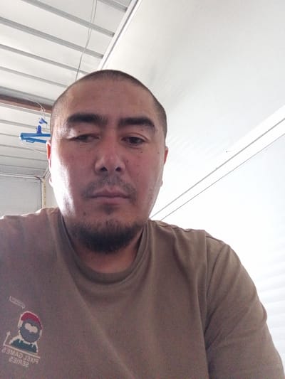 The image is a close-up selfie of a man with a shaved head and a goatee, wearing a tan t-shirt. The man is looking directly at the camera. The background appears to be an indoor space with a white ceiling and metal beams. On the man's t-shirt, there is a graphic with pixelated figures and text. The setting is likely a workshop or a similar utilitarian space. The image is taken from the chest up.