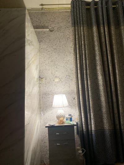 A room features textured wallpaper, a gray patterned curtain, and a marble countertop. A white lamp illuminates a bedside table. The table holds decorative items, including what looks like a small bouquet of flowers. A white cord runs from the lamp to an electrical outlet near the floor. The room is mostly gray and white with dim lighting.