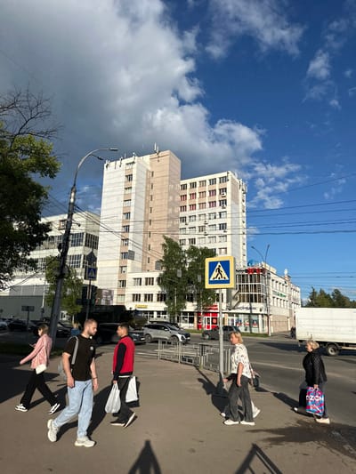 In Rubtsovo, Russia, on a bright day with a mix of clouds and blue sky, a wide street is bustling with activity. Pedestrians cross a street at a marked crosswalk in front of a large, modern multi-story building with a light-colored facade. Several people are walking, including a woman in a pink jacket, a man in a black t-shirt and light blue jeans carrying a backpack, and another man in a red and black jacket carrying white shopping bags. Two women are walking away from the viewer, one in a patterned shirt and dark pants, and the other in a black jacket carrying a colorful shopping bag. Cars and a white truck are visible on the street. The environment is urban, with power lines crisscrossing the sky and trees lining the sidewalk.