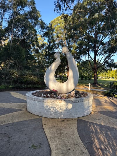 The image depicts a white stone sculpture of two abstract figures in an embrace, positioned within a circular, white-painted concrete planter. The planter contains dried leaves, small decorative items, and a few pink flowers. The sculpture and planter are situated on a paved area with a geometric pattern of grey and yellow concrete. Behind the sculpture, lush green trees and bushes form a natural backdrop, with a clear blue sky visible above. The scene appears to be outdoors, possibly in a park or garden setting. The lighting suggests it is either early morning or late afternoon, with long shadows cast across the ground. A bench is visible in the background to the right. This sculpture is located in the suburb of Chullora, Australia.