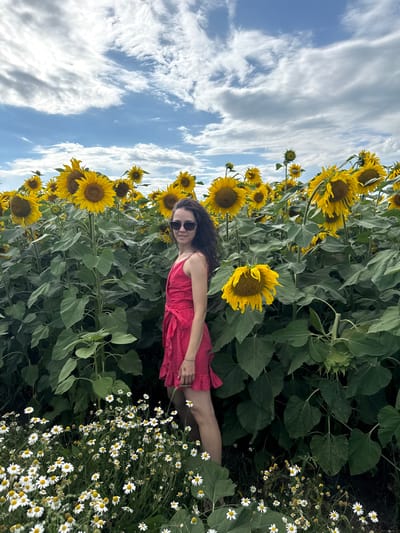 A young woman with long, dark, curly hair stands in a field of sunflowers and daisies under a cloudy sky. She is wearing a red, polka-dotted sundress and sunglasses. She is smiling and looking towards the camera. The sunflowers are tall and yellow, and the daisies are small and white. The sky is a mix of blue and white clouds. The image appears to be taken in a rural area, possibly on a farm or in the countryside. The location provided is Песчано-Коледино, Russia.