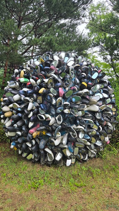 The image shows a large, spherical sculpture made of numerous car side mirrors. The sculpture is positioned outdoors, in front of lush green grass and behind it are trees, suggesting a natural or park-like setting. The mirrors are of various colors, including black, white, silver, red, blue, and yellow, and are clustered together to form the sphere. Some mirrors reflect the surrounding greenery and sky. The sculpture is located in the city of Gwangmyeong, South Korea, likely at a public art installation or park. There are no people or animals visible in the image.