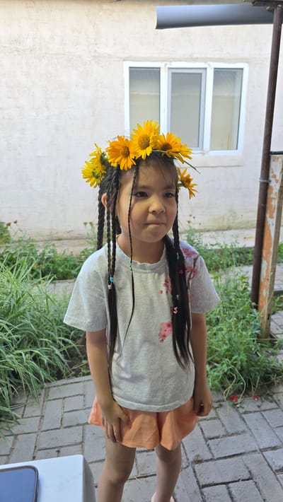 A young girl with braids adorned with a floral crown stands on a paved walkway in front of a weathered, light-colored building. She is wearing a light gray t-shirt with a subtle floral print and light orange shorts. Her braids, some with colorful beads, hang down her back and sides. The background features greenery, including tall grasses and some small red flowers, and a building with a white wall and a window. A wooden post is visible to the right of the girl. This scene is located in the city of Krugloozernoye, Kazakhstan.