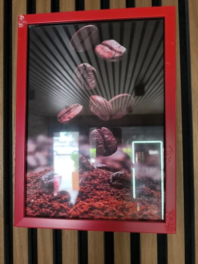 The image shows a framed picture of coffee beans. The frame is red and appears to be slightly damaged in the top left and bottom right corners, with some chipping and scuffing visible. The picture itself displays several large coffee beans suspended against a background of black lines radiating outwards, suggesting a dynamic or energetic theme. In the foreground of the picture, there's a pile of dark red coffee grounds with more coffee beans scattered on top. Through the glass of the frame, reflections are visible, indicating an indoor setting. These reflections include what appears to be a store interior with some lighting and possibly a white vehicle outside. The overall scene within the picture is focused on coffee imagery, likely intended for a cafe or a coffee-related business. The outer wall behind the frame is made of vertical wooden planks, alternating between a light wood color and black. There is no visible text in the image. The setting appears to be a commercial establishment, possibly a cafe, in Vulcănești, Moldova.