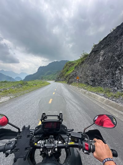 The image presents a first-person view from a motorcycle traveling on a winding mountain road under an overcast sky. The asphalt road, appearing damp, stretches forward with a yellow dashed center line. On the left, a rugged, dark gray rock face of a mountain rises steeply, sparsely covered with green vegetation. Further in the distance, layers of other mountains fade into the cloudy horizon.
In the foreground, parts of the motorcycle are visible, including the handlebar, windshield, and rearview mirrors. The rider's right hand, adorned with a blue and white patterned bracelet, is gripping the throttle. The reflection of the rider's red jacket can be seen in the right rearview mirror. The motorcycle's dashboard includes a digital display (showing "21:45" and other numbers) and a robust phone mount. A prominent component on the handlebar clamp bears the text "SPIRIT BEAST." Red buttons are visible on the motorcycle's right-hand control cluster.
A small yellow and red triangular road sign, indicating a left curve, is positioned near the base of the mountain face on the left side of the road. The gloomy, overcast weather creates a muted color palette dominated by grays and dark greens. This scene depicts a journey through the mountainous landscape of Minh Sơn Commune, Vietnam, highlighting the rugged terrain and an adventurous ride.