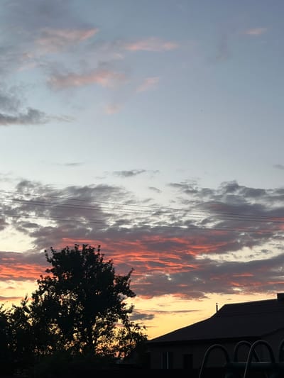 This is a photograph of a sunset sky with dramatic clouds in shades of pink, orange, and gray, over a silhouetted tree and the dark roof of a house in the distance. Power lines are visible horizontally across the middle of the frame. The location is the village of Sarotski, within the Sarotski Sel'sovet, Belarus. The image captures a natural scene with elements of rural infrastructure.