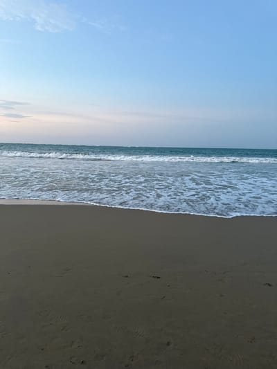 The image depicts a beach scene in Cabarete, Dominican Republic. The composition is vertically oriented, with the sky occupying the left portion of the frame and the beach and ocean on the right. The sky is a gradient of light blue, fading to a pale pink near the horizon. The ocean is visible as a strip with waves crashing onto the sand. The beach occupies the right side of the image and appears to be wet, with some footprints visible. No people are present in the frame.
