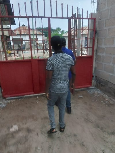 The image depicts a residential or semi-urban scene in Mwandege, Tanzania, viewed from an unpaved ground surface. Two individuals are shown from behind, walking away from the viewer towards a partially open, red metal gate. The person closer to the camera wears a black and white striped short-sleeved shirt, light grey trousers, and black sandals, with a gold-colored ring on their left hand. The second individual, partially obscured behind the first, wears a blue shirt and dark trousers with dark shoes.
The prominent red metal gate features solid lower panels and an upper section with vertical bars topped by finials. To the immediate right of the gate, an unfinished wall constructed of grey concrete blocks is visible. Beyond the gate, an unpaved area or street is visible, bordered by multiple single-story buildings. Several structures feature corrugated metal roofs, some showing signs of weathering. One building in the mid-background is white with a distinctive rounded or conical corrugated metal roof. Another building appears to be under construction or renovation, with scaffolding or timber supports near its front.
Further in the background, a person wearing yellow clothing is visible near a building on the left, possibly stooping or sitting. Another individual is walking towards the center-right background. Various forms of vegetation, including trees, are present beyond the buildings. A vertical utility pole and a dark sign with white lettering (partially visible, possibly "SAMHA") are discernible through the gate. The sky above is overcast or hazy, indicating an outdoor daytime setting.