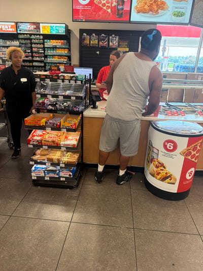 The image depicts the interior of a convenience store, likely a 7-Eleven, located in Hayti, Missouri, United States of America. A female employee, wearing a black uniform with a name tag reading "MAXIAM," stands behind a counter, interacting with a customer. The customer, a male wearing a grey tank top and grey shorts, is facing away from the camera, his back to the viewer. He has a blue hair pick in his hair. Various merchandise, including candy bars, packaged snacks, and drinks, are displayed on shelves and in refrigerated units throughout the store. A large promotional graphic for breakfast burritos and pizza is visible on a cylindrical display near the customer. The store appears to be well-lit and stocked.