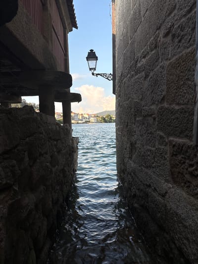 The image shows a narrow waterway between two stone buildings, with the sea visible in the background. On the left, a building with wooden siding and a red roof is supported by stone pillars. A lamppost with an ornate bracket is mounted on the wall of the building on the right, which is constructed of rough-hewn stone blocks. The water is choppy and reflects the sunlight. In the distance, across the water, is a town with buildings and a green hillside. The scene is located in the town of Combarro, Spain, a coastal village known for its traditional architecture and seafood restaurants. The time of day appears to be late afternoon or early evening, as indicated by the warm sunlight and the shadows.