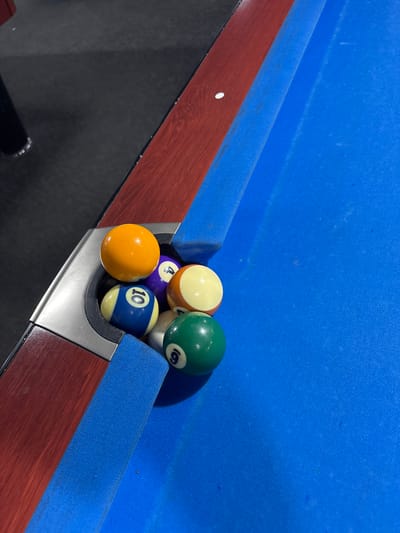 This image captures a close-up, angled view of a corner pocket on a blue-felted pool table, located in Arnhem, Netherlands. Six billiard balls are collected within and immediately around the metallic, silver-toned pocket with its black rubber lining.
The table features a vibrant blue felt playing surface, which exhibits minor signs of wear, dust specks, and slight discoloration. The table rails are constructed from a reddish-brown wood with a distinct grain pattern, separated from the felt by a thin dark trim.
The visible billiard balls include:
* A solid orange ball, positioned highest on the edge of the pocket.
* A solid purple ball marked with the number '4'.
* A striped yellow and tan/off-white ball, partially obscured.
* A striped blue and white ball clearly marked with the number '10'.
* A solid green ball marked with the number '9'.
* A partially visible white cue ball nestled amongst the others.
The background to the upper left of the table displays a dark, matte floor or surface, with a dark cylindrical object, possibly a table leg, visible in the far upper-left corner. The scene is illuminated by what appears to be ambient or overhead artificial lighting. No individuals, actions, or interactions are visible within the frame.