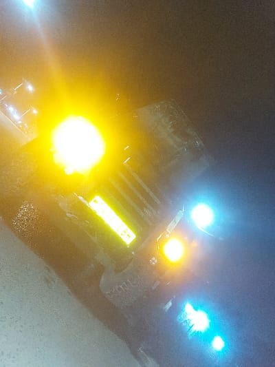 The scene is captured at night in Msata, Tanzania. A large vehicle, likely a truck, is positioned diagonally across the frame, its form largely obscured by intense light sources. The vehicle displays a dominant, bright yellow-white light in the upper left-center. A rectangular yellow reflective object, possibly a license plate, is visible below this light, showing partial characters "TBH." To the right, two distinct yellow circular lights and two distinct blue-cyan circular lights are illuminated. The ground surface in the foreground appears dark and reflective, suggesting wetness or a polished surface. No individuals are visible, and no specific actions or interactions are discernible.