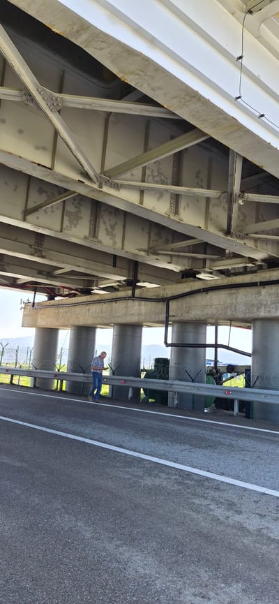 A man in blue jeans and a light-colored plaid shirt stands on a paved road beneath a large concrete and steel bridge structure. He is holding a mobile device in his right hand and looking down at it. The man is positioned to the left of the frame, facing right. Behind him, a metal guardrail with barbed wire topping lines the edge of the road. Beyond the guardrail are several large, cylindrical concrete pillars supporting the bridge. Further back, a green cylindrical structure, possibly a tank, is visible, along with fencing and sparse vegetation. The sky above is clear and bright. The environment appears to be an outdoor, possibly industrial or transportation-related area, likely on the outskirts of Volno-Nadejdinskoe, Russia, given the prompt's context. The bridge's underside is a complex network of steel beams and supports.