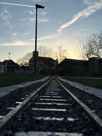 The image presents a low-angle view of a miniature railway track extending into the distance in Olingen, Luxembourg, under a late afternoon or early morning sky. The track, composed of metal rails on dark grey gravel ballast with regularly spaced wooden sleepers, is flanked by light grey concrete edgings and short green grass. In the midground, a tall, dark street light pole stands prominently to the left of the tracks. Further back on the left, a smaller, dark lamp post is visible beyond some metallic structures, possibly fencing or playground equipment. The background features several buildings with dark roofs and light-colored walls, partially obscured by a dense arrangement of bare deciduous trees and shrubs. One building, centrally located, exhibits a distinctive rounded or turret-like roof section. On the far right, a rectangular green utility box is visible. The sky transitions from a pale blue at the zenith to a soft yellowish-orange glow at the horizon on the right, indicating a low sun. Wispy white clouds are scattered across the sky, with a prominent, long contrail stretching diagonally from the upper left. No individuals or active subjects are present in the scene.