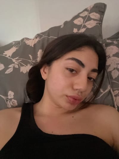 This is a slightly blurry, close-up, selfie-style image taken indoors, likely in a bedroom. The subject is a young woman with dark hair, lying on her side on a pillow with a floral pattern. She is looking towards the camera with her lips slightly parted. She is wearing a black tank top. The lighting appears to be artificial, suggesting it could be evening or nighttime, or an indoor setting with dim lighting. The overall mood is relaxed and intimate. There is no visible text in the image.