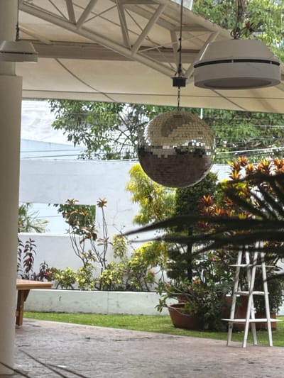 The image, originally rotated 90 degrees clockwise, captures an outdoor or semi-outdoor setting, possibly a patio, garden, or recreational area, under natural daylight.
In the upper left quadrant of the upright image, a white, cylindrical object resembling a speaker or light fixture is suspended from a white overhead structure, likely part of an awning or pergola. Below and slightly to the right of this object, a large, spherical disco ball, covered in numerous small, reflective silver squares, hangs by a chain.
The background is dominated by a rich array of green foliage, including dense trees and bushes, some displaying autumnal or variegated colors with yellowish and reddish leaves. Behind the greenery, parts of a modern, white building or wall are visible. Towards the right side of the frame, a decorative white fence, possibly made of metal or wood, stands amidst the plants, featuring pointed tops on its vertical elements. Beyond the fence, a section of green grass is visible, bordering a grey, paved pathway or concrete surface that runs along the far right edge of the image.
The scene is devoid of people or explicit activity, presenting a tranquil ambiance. The presence of the disco ball suggests the space is used for gatherings or celebrations. The lighting indicates it is daytime. No visible text is present in the media file. The context for the location is Tres de Mayo, Mexico.