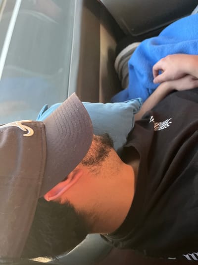 The image depicts two individuals inside a vehicle, located in the city of Leiria, Portugal. The perspective is an overhead view, with the subjects positioned horizontally across the frame.
In the foreground and occupying the lower-left portion of the image, a person is observed resting their head on a light blue fabric cushion. This individual wears a dark baseball cap with a light-colored emblem on the front, resembling a crescent or the number '5'. They are dressed in a black t-shirt with a white graphic logo visible on the right chest area. Dark facial hair is present on their jawline and neck. Their visible skin exhibits a reddish-brown tone, potentially from ambient light.
In the upper-right section of the image, the left arm and partially clasped hand of a second individual are visible. This hand rests upon a bright blue blanket.
The background consists of a light grey panel, possibly an interior wall or window frame, and darker upholstered surfaces, consistent with the interior of a vehicle.