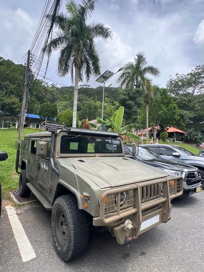 The media file displays a military-style vehicle, likely a Humvee, parked on the side of a road. The vehicle is olive green and features a reinforced bumper and a light bar on the roof. A dark-colored SUV is parked behind it. The scene is set in a tropical environment with lush green trees and palm trees visible in the background. A hill covered in foliage rises behind the trees. Power lines and a lamppost are present to the left of the vehicles. The sky is overcast with clouds. A sign on the side of the Humvee, visible in the lower left portion of the image, contains Thai script and English text identifying it as the "KHUANDON IMMIGRATION CHECK POINT SATUN IMMIGRATION." There are small flags planted in the ground in the middle ground, suggesting a border crossing or official checkpoint. The overall impression is one of a functional, possibly remote, location. The weather appears to be overcast, and the time of day is difficult to ascertain precisely but seems to be daytime. There are no people clearly visible.