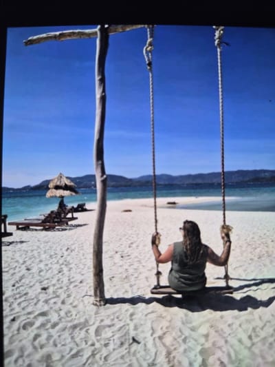 The image shows a person swinging on a rope swing on a sandy beach. The background includes a blue sky and mountains. There is no evidence to suggest that this image was taken in Mont-Saint-Martin, France.