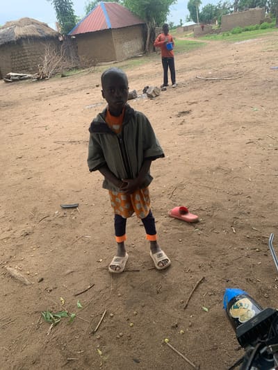 A young boy stands in the foreground of the image, looking directly at the camera. He is wearing a grey hooded jacket over an orange shirt and orange shorts with a repeating elephant pattern. His legs are covered by dark blue leggings that end in bright orange cuffs. He wears a single white sandal on his left foot. His right foot is bare. The ground is dry, sandy earth with scattered leaves and twigs.

In the background, two traditional huts are visible. The hut on the left has a thatched roof, while the hut on the right has a red metal roof with blue trim. A second, older boy or young man is standing further back, on the right side of the frame, holding a blue bucket and looking to his left. Beyond the huts, more buildings and trees can be seen under a cloudy sky. The scene appears to be a rural village in Dull A, Nigeria.