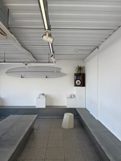 This indoor shot depicts an asymmetrical interior space. The room is characterized by differing wall treatments: on the left, a wall of corrugated metal, and on the right, a section of grey stone tilework. The lighting fixtures are attached to a metal track.

A surfboard, mounted vertically, adorns the middle wall. A vintage speaker and a potted plant are placed on a shelf near the top. A small, white cube sits next to the speaker, holding papers. Another, similar cube sits lower on the wall. On the stone-tiled side, a white, geometric sculpture is partially visible.

This image was taken in Toseong-myeon, South Korea.